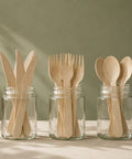 Set of wooden cutlery including knives, forks, and spoons in clear jars on a neutral background.