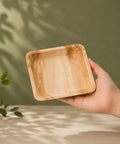 Hand holding a wooden square plate against a green background with leaves.
